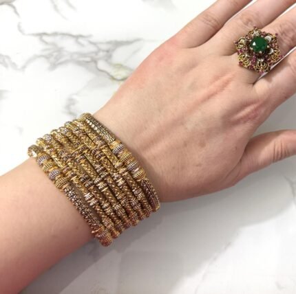 A hand wearing eight intricately detailed gold-tone bangles with dual-tone embellishments and a statement green gemstone ring, displayed on a white marble surface.