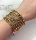 A hand adorned with a set of 8 antique gold bangles, featuring intricate floral motifs and embedded clear crystals, resting on a marble surface.