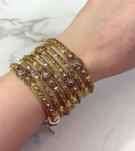 A hand adorned with a set of 8 antique gold bangles, featuring intricate floral motifs and embedded clear crystals, resting on a marble surface.