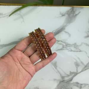 A hand holding a pair of antique gold bangles featuring intricate floral patterns with a dual-tone finish, displayed on a marble background.