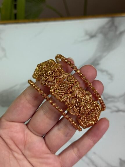 A wide gold kada bangle with intricate floral engravings and beadwork, held in a hand against a marble background.