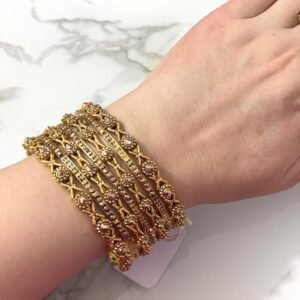 A hand adorned with eight intricately designed antique gold bangles featuring cross motifs and embedded pearl-like beads, displayed on a marble surface.