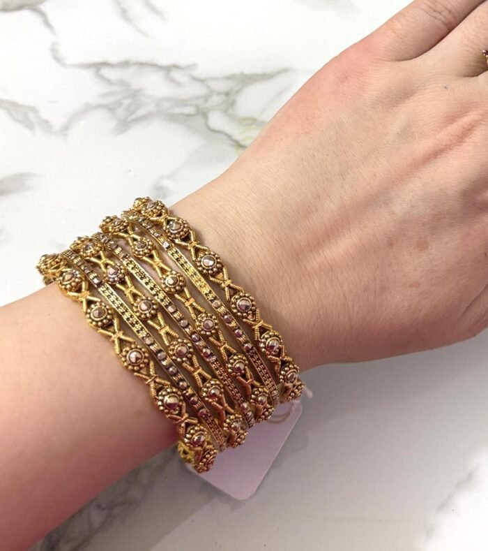 A hand adorned with eight intricately designed antique gold bangles featuring cross motifs and embedded pearl-like beads, displayed on a marble surface.
