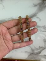 A close-up of a hand holding a pair of gold-plated ethnic bangles with intricate filigree patterns and ruby red stone embellishments, against a marble background.