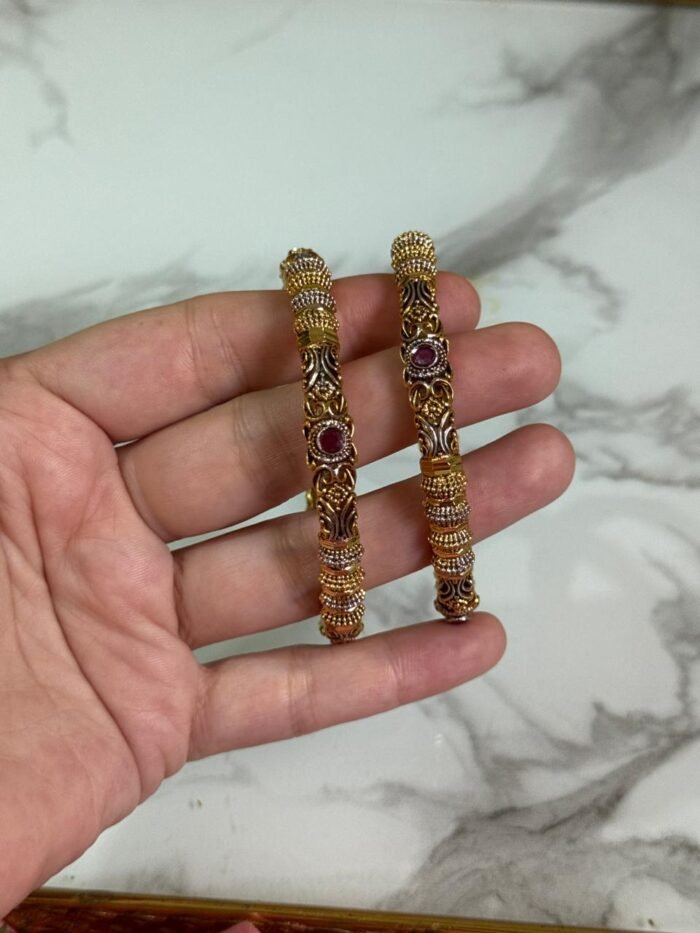 A close-up of a hand holding a pair of gold-plated ethnic bangles with intricate filigree patterns and ruby red stone embellishments, against a marble background.