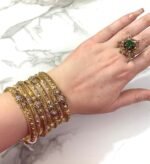 A hand adorned with a set of 8 antique gold bangles, featuring intricate floral motifs and embedded clear crystals, resting on a marble surface.