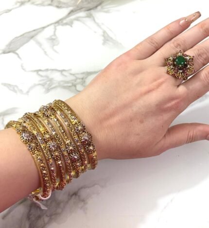 A hand adorned with a set of 8 antique gold bangles, featuring intricate floral motifs and embedded clear crystals, resting on a marble surface.