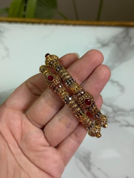 A close-up of a hand holding a pair of ornate gold-plated bangles, featuring maroon centerpiece stones and intricate black enamel accents, displayed against a marble background.