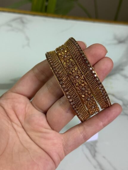 A hand holding a set of three gold bangles, each encrusted with detailed crystal patterns in symmetrical and wave designs, displayed against a marble background.