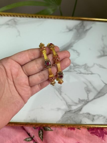 A hand holding two traditional-style gold bangles featuring crown-like ends with red gemstone embellishments and intricate detailing, set against a marble background.