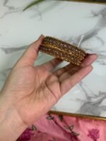 A hand holding a pair of antique gold bangles featuring intricate floral patterns with a dual-tone finish, displayed on a marble background.