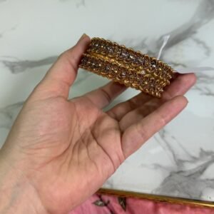 A hand holding a pair of antique gold bangles featuring intricate floral patterns with a dual-tone finish, displayed on a marble background.