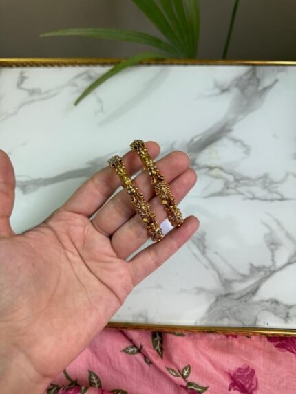 A hand holding two intricately crafted gold bangles featuring antique-style filigree work and floral motifs, with a marble surface in the background.