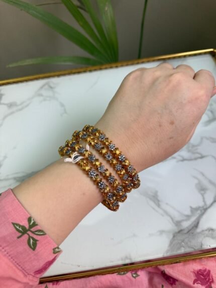 A set of 4 gold and silver dual-tone bangles adorned with alternating floral motifs, worn on a wrist over a pink floral sleeve, placed against a white marble background with a green plant in the frame.