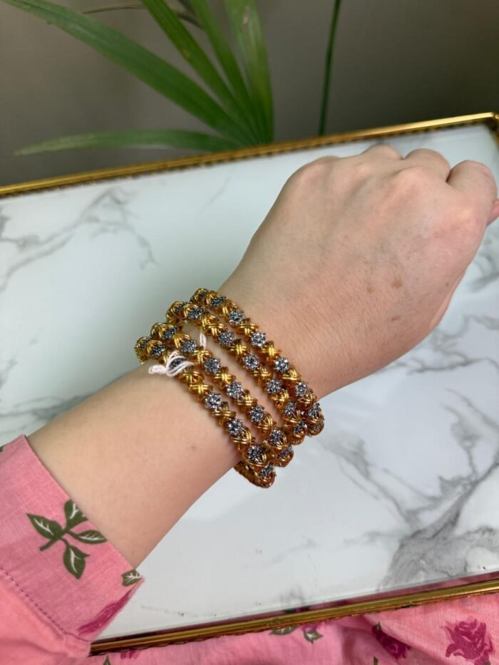 A set of 4 gold and silver dual-tone bangles adorned with alternating floral motifs, worn on a wrist over a pink floral sleeve, placed against a white marble background with a green plant in the frame.