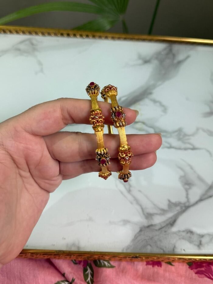 A hand holding two traditional-style gold bangles featuring crown-like ends with red gemstone embellishments and intricate detailing, set against a marble background.