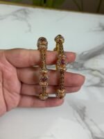 A pair of ornate gold bangles adorned with clusters of multicolor stones in shades of red, pink, and white, held in a hand over a marble background.