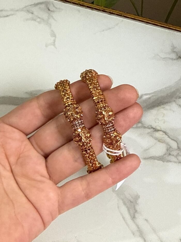 A pair of traditional gold bangles with rope-texture design and ornate bead detailing, held in hand over a marble surface.