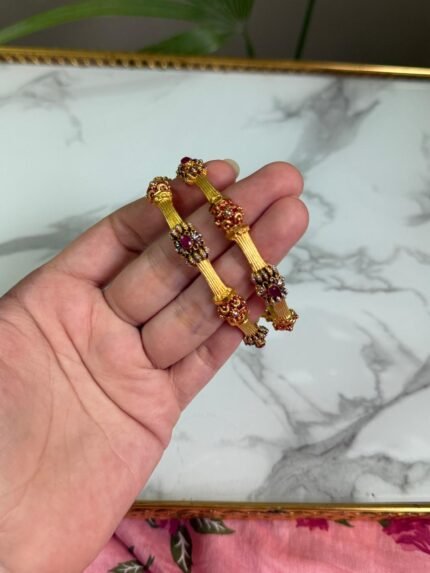 A hand holding two traditional-style gold bangles featuring crown-like ends with red gemstone embellishments and intricate detailing, set against a marble background.