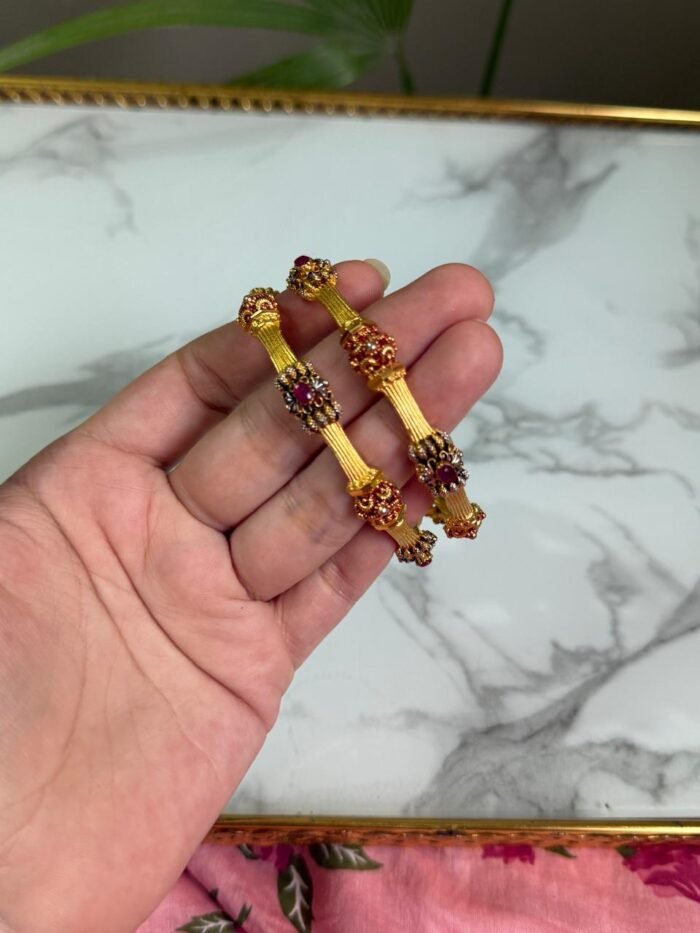 A hand holding two traditional-style gold bangles featuring crown-like ends with red gemstone embellishments and intricate detailing, set against a marble background.