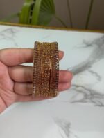 A hand holding a set of three gold bangles, each encrusted with detailed crystal patterns in symmetrical and wave designs, displayed against a marble background.