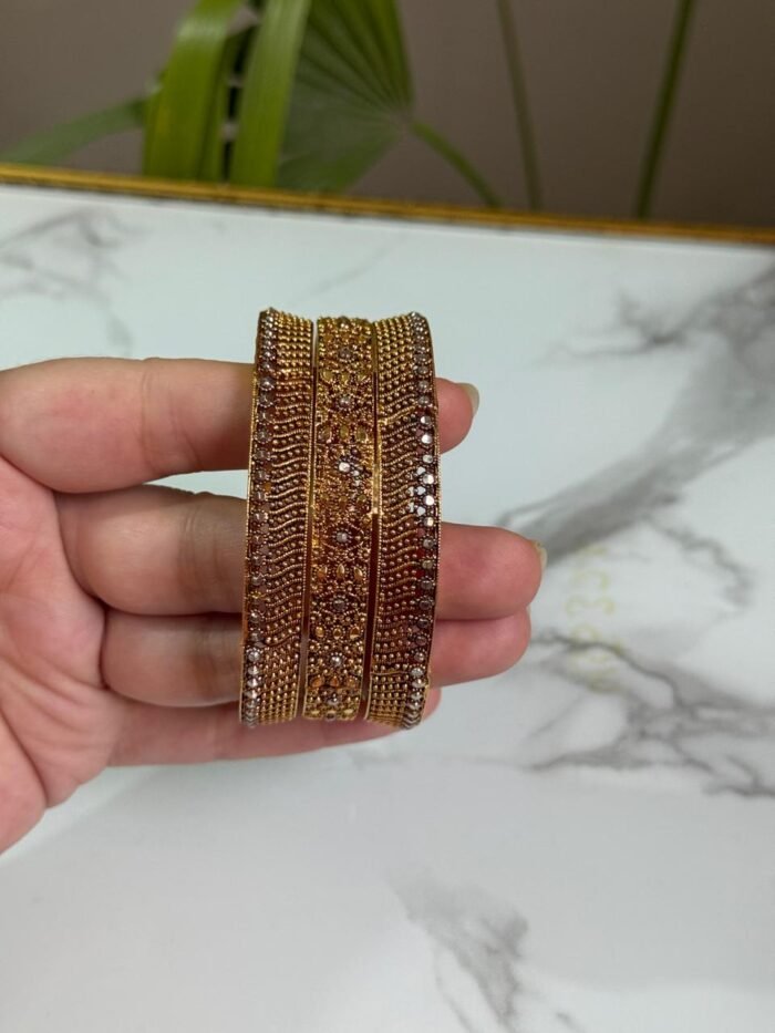 A hand holding a set of three gold bangles, each encrusted with detailed crystal patterns in symmetrical and wave designs, displayed against a marble background.