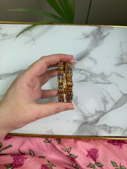A hand holding a pair of antique gold-toned bangles with intricate floral filigree work and beaded textures, placed against a marble background with a touch of greenery.