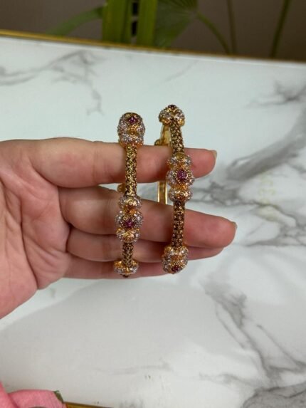 A pair of ornate gold bangles adorned with clusters of multicolor stones in shades of red, pink, and white, held in a hand over a marble background.