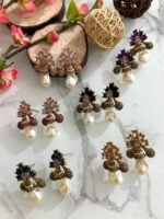 A collection of floral gemstone and pearl drop earrings in multiple colors, including pink, purple, blue, black, and gold, displayed on a white marble surface with decorative flowers and wooden props.