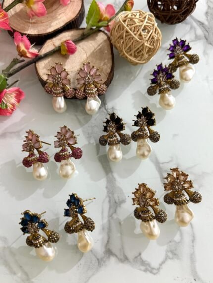A collection of floral gemstone and pearl drop earrings in multiple colors, including pink, purple, blue, black, and gold, displayed on a white marble surface with decorative flowers and wooden props.