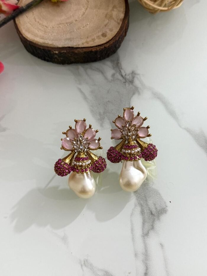 A collection of floral gemstone and pearl drop earrings in multiple colors, including pink, purple, blue, black, and gold, displayed on a white marble surface with decorative flowers and wooden props.