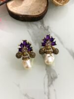 A collection of floral gemstone and pearl drop earrings in multiple colors, including pink, purple, blue, black, and gold, displayed on a white marble surface with decorative flowers and wooden props.