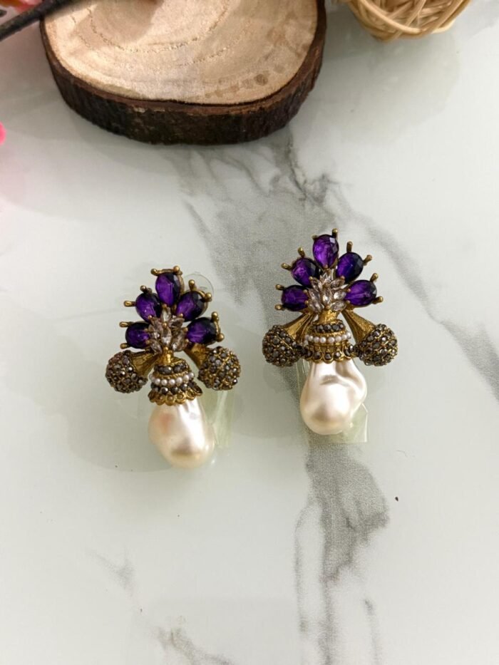 A collection of floral gemstone and pearl drop earrings in multiple colors, including pink, purple, blue, black, and gold, displayed on a white marble surface with decorative flowers and wooden props.