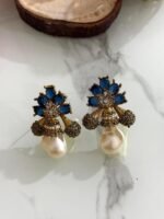 A collection of floral gemstone and pearl drop earrings in multiple colors, including pink, purple, blue, black, and gold, displayed on a white marble surface with decorative flowers and wooden props.