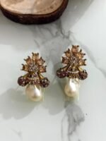 A collection of floral gemstone and pearl drop earrings in multiple colors, including pink, purple, blue, black, and gold, displayed on a white marble surface with decorative flowers and wooden props.