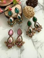 A set of traditional teardrop earrings in green and pink with kundan-inspired detailing, pearl inlays, and floral metalwork, displayed on a marble surface with wooden and floral props.