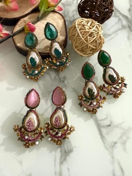 A set of traditional teardrop earrings in green and pink with kundan-inspired detailing, pearl inlays, and floral metalwork, displayed on a marble surface with wooden and floral props.