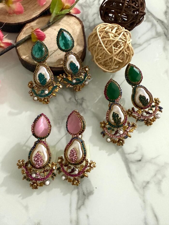 A set of traditional teardrop earrings in green and pink with kundan-inspired detailing, pearl inlays, and floral metalwork, displayed on a marble surface with wooden and floral props.