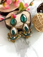 A set of traditional teardrop earrings in green and pink with kundan-inspired detailing, pearl inlays, and floral metalwork, displayed on a marble surface with wooden and floral props.