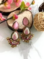 A set of traditional teardrop earrings in green and pink with kundan-inspired detailing, pearl inlays, and floral metalwork, displayed on a marble surface with wooden and floral props.