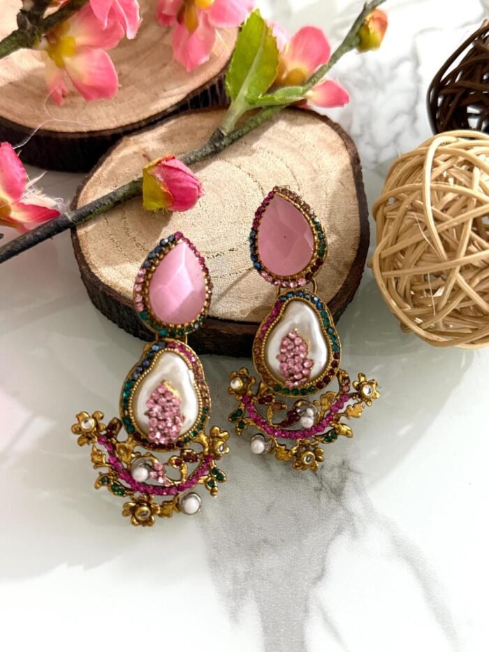 A set of traditional teardrop earrings in green and pink with kundan-inspired detailing, pearl inlays, and floral metalwork, displayed on a marble surface with wooden and floral props.