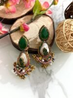 A set of traditional teardrop earrings in green and pink with kundan-inspired detailing, pearl inlays, and floral metalwork, displayed on a marble surface with wooden and floral props.