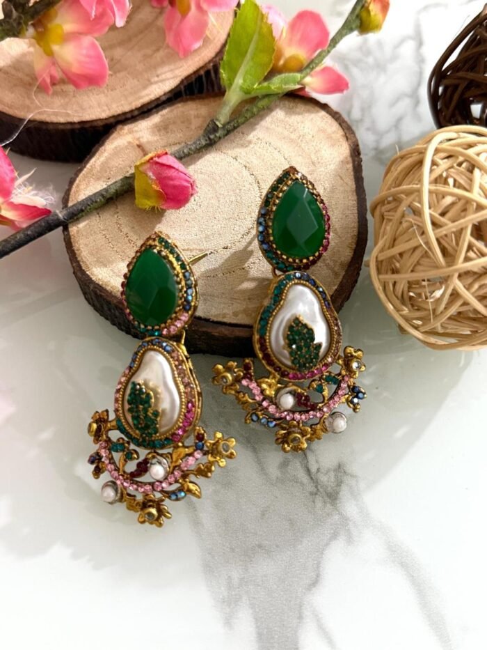 A set of traditional teardrop earrings in green and pink with kundan-inspired detailing, pearl inlays, and floral metalwork, displayed on a marble surface with wooden and floral props.