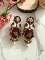 Four pairs of ornate earrings with red and black stones, pearl drops, and gold detailing, placed on a marble surface with floral and wooden decorative props in the background.