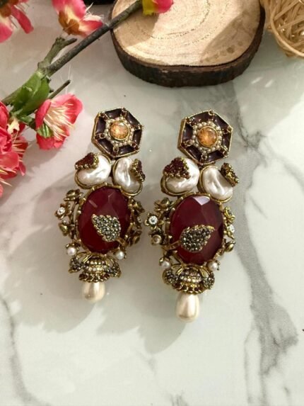 Four pairs of ornate earrings with red and black stones, pearl drops, and gold detailing, placed on a marble surface with floral and wooden decorative props in the background.