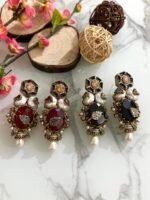 Four pairs of ornate earrings with red and black stones, pearl drops, and gold detailing, placed on a marble surface with floral and wooden decorative props in the background.