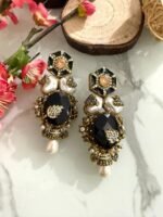 Four pairs of ornate earrings with red and black stones, pearl drops, and gold detailing, placed on a marble surface with floral and wooden decorative props in the background.