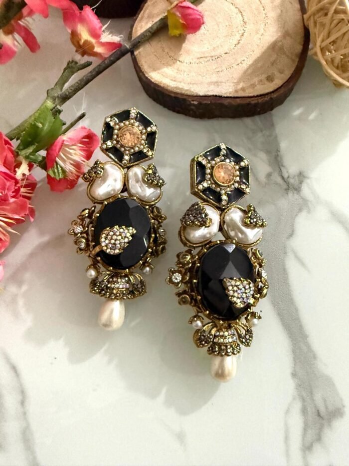 Four pairs of ornate earrings with red and black stones, pearl drops, and gold detailing, placed on a marble surface with floral and wooden decorative props in the background.