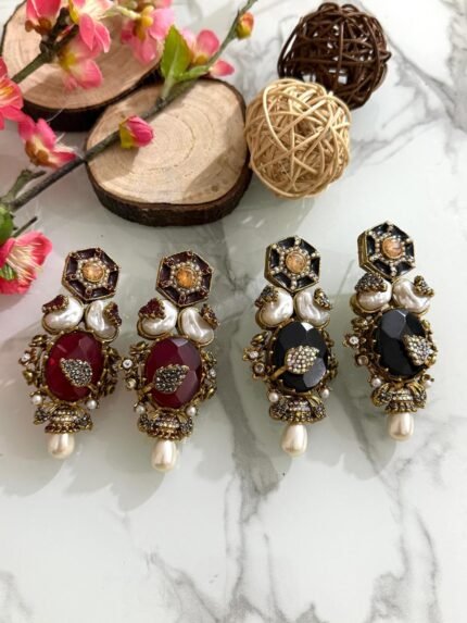 Four pairs of ornate earrings with red and black stones, pearl drops, and gold detailing, placed on a marble surface with floral and wooden decorative props in the background.