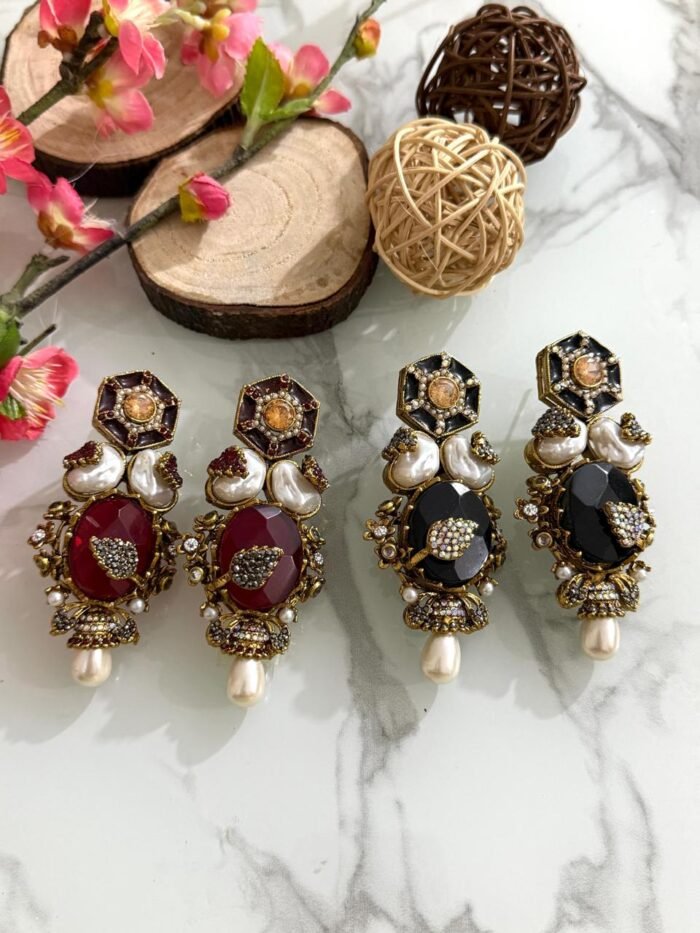 Four pairs of ornate earrings with red and black stones, pearl drops, and gold detailing, placed on a marble surface with floral and wooden decorative props in the background.